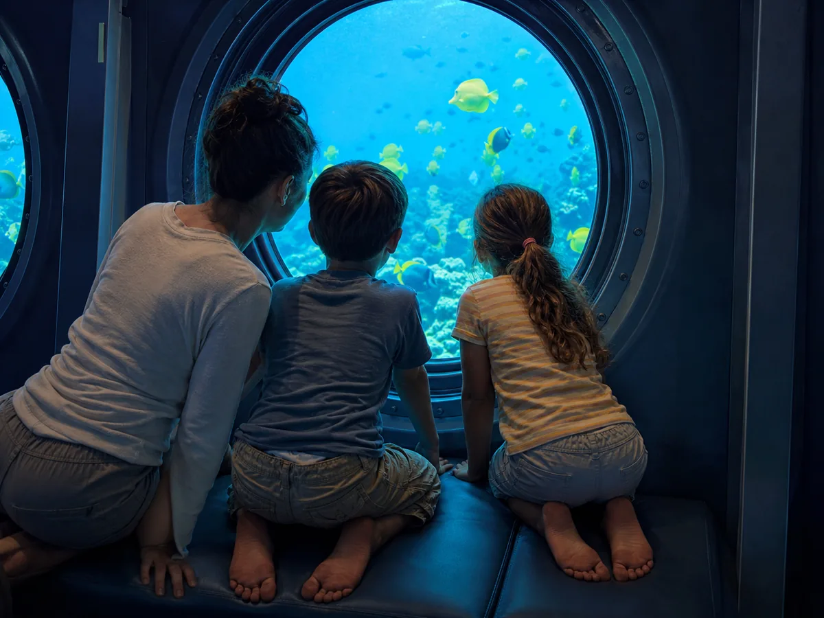 Children and a parent looking out a round submarine porthole at blue water and yellow fish