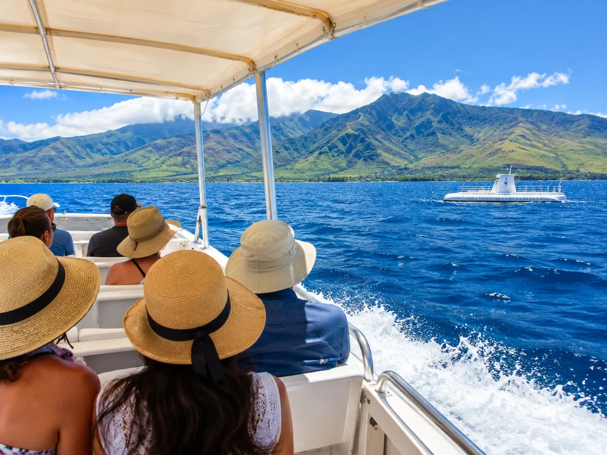 An open-air shuttle boat crossing blue water toward a white submarine waiting on the surface